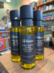 Three bottles of Kiwi Artisan Black Truffle Infused Extra Virgin Olive Oil on a wooden surface with a store shelf in the background.