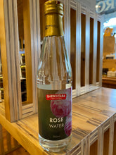 A glass bottle of Sheikhtaba Rose Water, with a label showing the product name and a picture of a rose. The bottle is placed on a wooden surface.